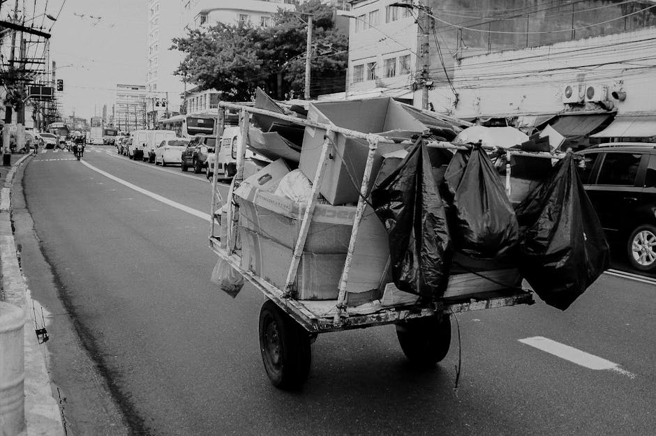 A small, wheeled cart loaded with various discarded items, including cardboard boxes, black plastic rubbish bags, and other miscellaneous debris, moving along a city street. The cart appears to be made of metal with visible wear and rust, supporting a bulky, uneven collection of waste materials. The cardboard boxes are partially open, showing rough, corrugated textures, while the black rubbish bags are tied at the top, hanging loosely over the sides of the cart. Surrounding the cart, the street is lined with parked vehicles on both sides, mainly modern cars, with a mixture of sedans and SUVs, and a few pedestrians visible further down the road. In the background, multi-storey buildings with window air conditioning units and electrical wiring add to the urban environment. The scene is captured during daylight hours, with natural, diffused lighting highlighting the contrast between the various materials and textures, subtly relating to the context of private waste handling and alternative rubbish clearance services provided by Rubbish Clearance Stockwell.