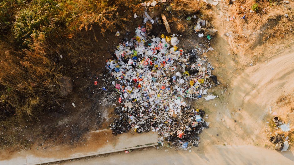 An aerial view of an uncovered waste dump on an outdoor site showing a large pile of mixed rubbish, primarily composed of plastic bottles, bags, and packaging materials, scattered across a patch of bare, uneven ground with dirt and small rocks. The pile is situated near the edge of a construction or excavation site, with a section of concrete pathway visible at the bottom of the image. Surrounding the refuse are areas of sparse, dry vegetation on the left side, indicating minimal ground cover. The scene is illuminated by natural daylight, casting soft shadows from the accumulated waste. This setting illustrates a location where private or independent rubbish collection may be required, highlighting the importance of proper waste management and clearance services provided by companies like Rubbish Clearance Stockwell targeting misplaced or illegally dumped rubbish in outdoor environments.