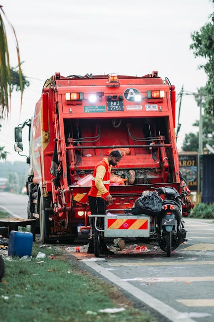 A small, wheeled cart loaded with various discarded items, including cardboard boxes, black plastic rubbish bags, and other miscellaneous debris, moving along a city street. The cart appears to be made of metal with visible wear and rust, supporting a bulky, uneven collection of waste materials. The cardboard boxes are partially open, showing rough, corrugated textures, while the black rubbish bags are tied at the top, hanging loosely over the sides of the cart. Surrounding the cart, the street is lined with parked vehicles on both sides, mainly modern cars, with a mixture of sedans and SUVs, and a few pedestrians visible further down the road. In the background, multi-storey buildings with window air conditioning units and electrical wiring add to the urban environment. The scene is captured during daylight hours, with natural, diffused lighting highlighting the contrast between the various materials and textures, subtly relating to the context of private waste handling and alternative rubbish clearance services provided by Rubbish Clearance Stockwell.