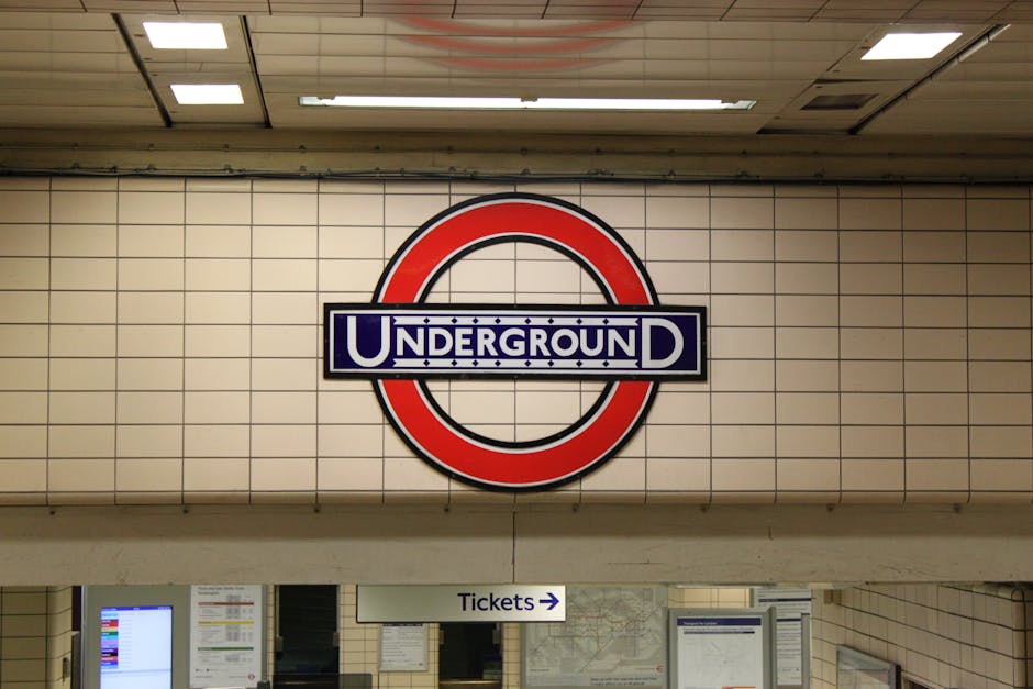 A large, circular London Underground sign featuring a red outer ring and a blue rectangular bar with the word 'UNDERGROUND' in white capital letters, mounted on a tiled white wall inside an underground station. Below the sign, there is a white sign with black text indicating directions to ticket areas, with an arrow pointing to the right. The environment is well-lit with fluorescent ceiling lights, and the tiles have a glossy finish. The station appears clean and organized, representing typical London's transit infrastructure connected to urban waste and rubbish management within the station environment, near facilities such as ticket counters and information screens.