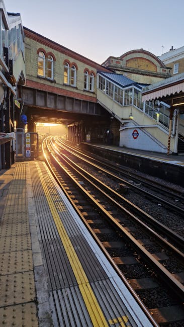 A view of Stockwell Tube Station platform during daylight hours, showing the railway tracks curving into the distance under the station bridge. The platform features tactile yellow warning strips along the edge and textured yellow lines for safety. On the right side, there is a white staircase with a metal railing leading up to an overpass, which is attached to the station building constructed from brick and metal elements. The station building has arched windows with a brick facade, and the elevated walkway area includes glass-enclosed sections. In the background, sunlight filters through the opening under the bridge, casting a warm glow on the tracks and platform surface. The environment is clean and well-maintained, illustrating a typical urban underground station setting, with no visible rubbish or debris. This scene naturally relates to the importance of maintaining clear, safe station environments, an aspect legal and independent waste handling services like Rubbish Clearance Stockwell can support by providing rubbish removal solutions to keep such public spaces tidy and accessible.