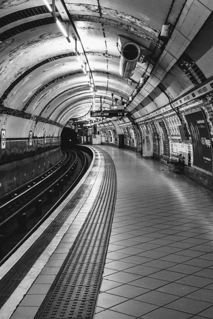 A large, circular London Underground sign featuring a red outer ring and a blue rectangular bar with the word 'UNDERGROUND' in white capital letters, mounted on a tiled white wall inside an underground station. Below the sign, there is a white sign with black text indicating directions to ticket areas, with an arrow pointing to the right. The environment is well-lit with fluorescent ceiling lights, and the tiles have a glossy finish. The station appears clean and organized, representing typical London's transit infrastructure connected to urban waste and rubbish management within the station environment, near facilities such as ticket counters and information screens.