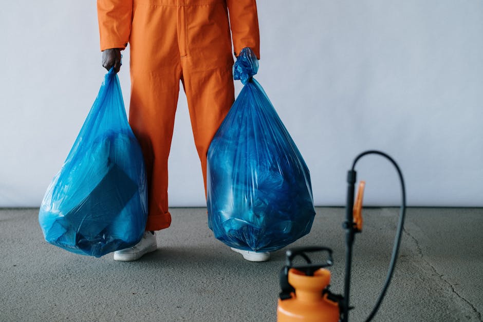 In the image, a person is standing indoors on a plain, light-colored floor, holding two large blue plastic rubbish bags filled with waste, one in each hand. The individual is wearing orange work overalls and white footwear, with their hands covered in dark gloves. The background is a neutral, light grey wall. In the foreground, partially visible, is an orange portable vacuum or extraction device connected to a black hose, suggesting a waste collection or cleaning process. The scene appears to be part of an internal waste management or rubbish removal activity, possibly undertaken by a professional service like Rubbish Clearance Stockwell, which specialises in private rubbish collection, on-site clearance, or alternative waste handling solutions. The focus on the filled rubbish bags and associated equipment indicates a structured rubbish removal operation aiming for efficient waste disposal within a controlled environment, aligning with the context of independent waste clearance services for residential or commercial properties.