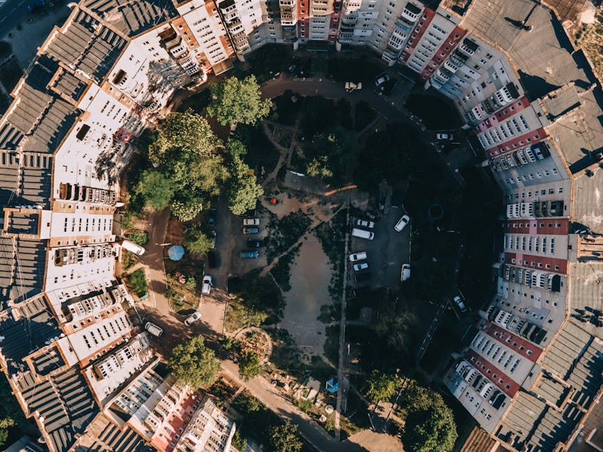 An aerial view of a circular residential courtyard surrounded by multi-storey terraced houses with pitched roofs, featuring a central green space with several mature trees, pathways, and a small playground area. The courtyard contains parked cars along the perimeter, with some on driveways and others on the paved road. The buildings are painted in light colors with red, beige, and white facades, and the rooftops are tiled in dark shades. Shadows cast by the buildings and trees suggest a sunny day. The scene illustrates a typical urban environment where private waste collection services, such as those from Rubbish Clearance Stockwell, may be employed for managing rubbish and waste disposal in place of municipal collection, with the dense arrangement of housing and designated parking areas surrounding the green communal space.